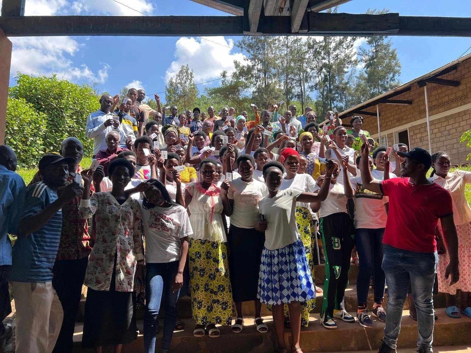 A large crowd of men, women and children standing together on a grassy slope outside a brick building, many with their hands raised.