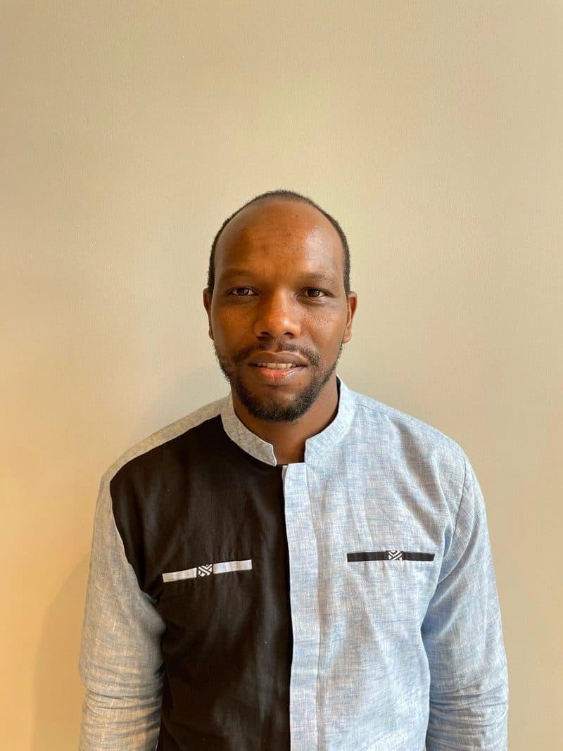 Portrait of Aimable Mugabo wearing a cream and black collarless shirt against a plain wall.