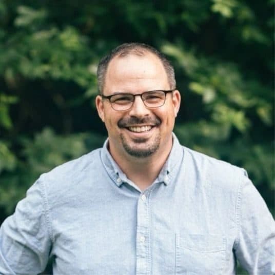 Portrait of Dave Shockey in a light blue shirt and glasses, photographed outdoors in front of foliage.