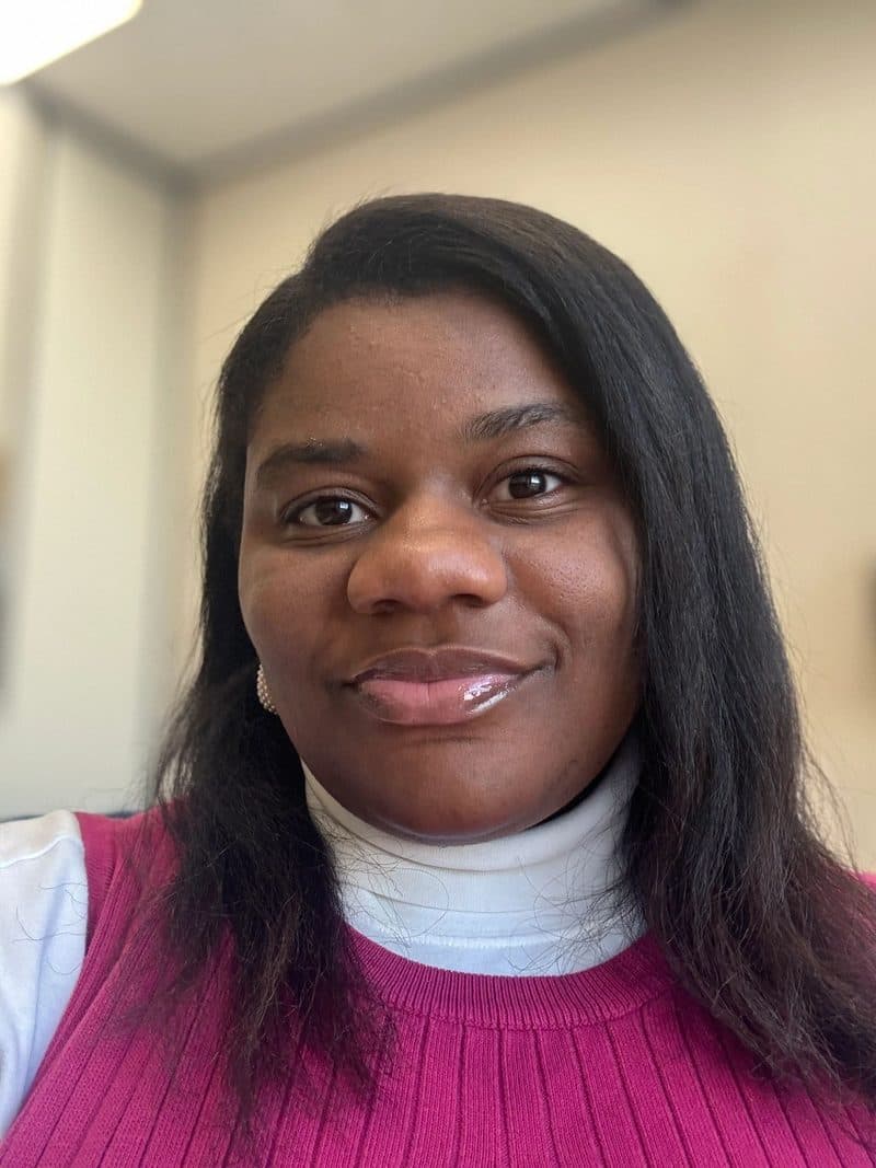 Portrait of Faith Jones smiling, wearing a white polo neck under a bright pink knitted vest.