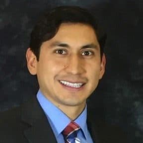 Portrait of Isaac Conde in a dark suit, blue shirt and patterned tie against a dark background.